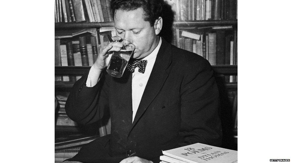 Welsh poet Dylan Thomas (1914 - 1953) drinking a glass of beer and smoking while seated at a desk with stacks of his books of poetry, New York City, c. 1950.