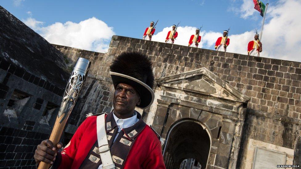 A man in an old-fashioned uniform holds the Queen's Baton. A row of men in similar clothing stand to attention behind him.