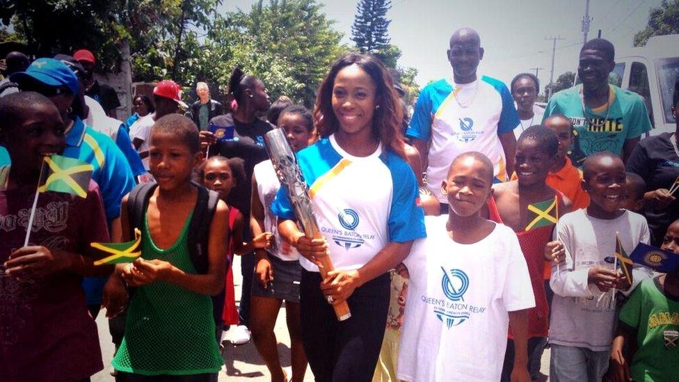 Female athlete holds the Queen's Baton, surrounded by smiling children waving Jamaican flags.