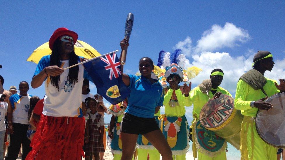 Woman holds Queen's Baton aloft as a carnival band play in the background.