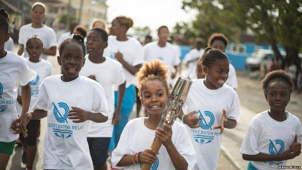 A group of children running together. A young girl holds the Queen's Baton.