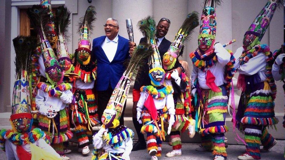 Two formally-dressed men hold the Queen's Baton, surrounded by a group of carnival dancers.