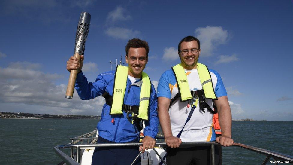 Two men wearing lifejackets pose with the Queen's Baton on board a small boat.