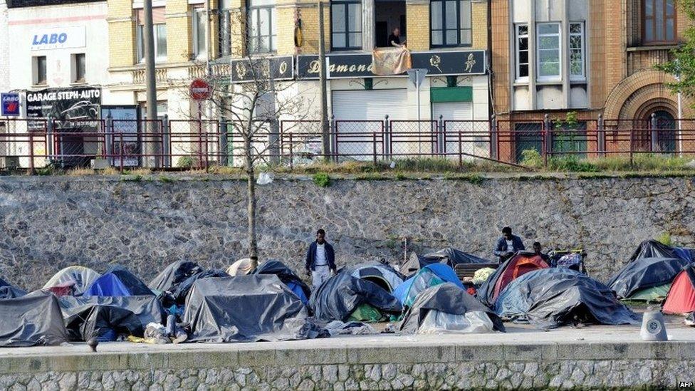 Migrants stand next to tents at their makeshift camp set up on a bank in Calais