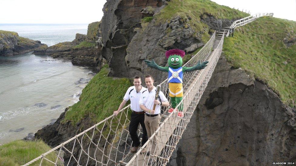 Two men pose with the Queen's Baton on a narrow rope bridge accompanied by a person dressed as a cartoon thistle.