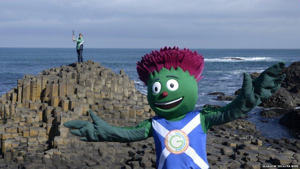 Woman standing on a coastal hexagonal rock formation holds Queen's Baton aloft while a person dressed as a cartoon thistle waves in the foreground.