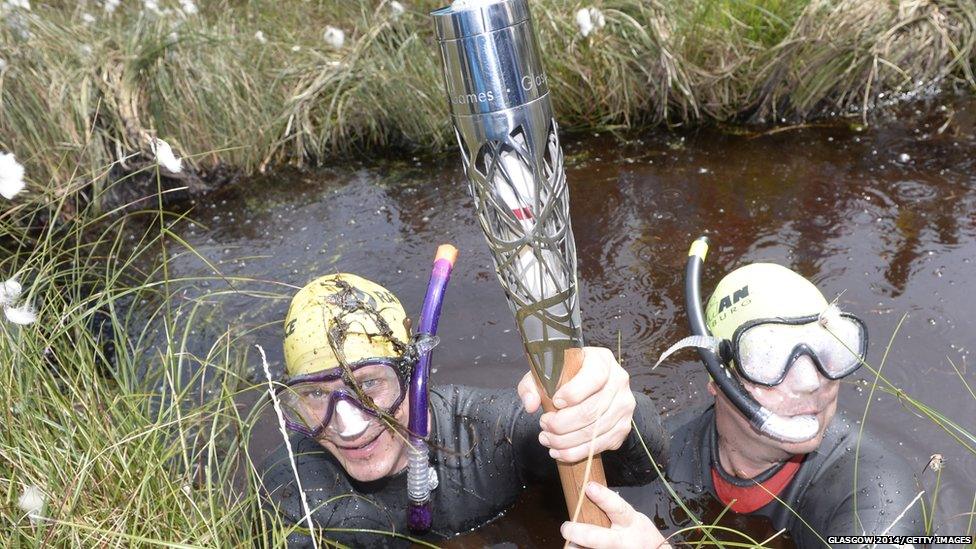 Two men wearing wetsuits and snorkelling equipment hold the Queen's Baton clear of the peat bog they are almost submerged in.