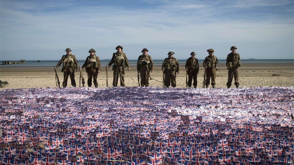 People in WW2 uniforms standing behind many small union flags on a beach