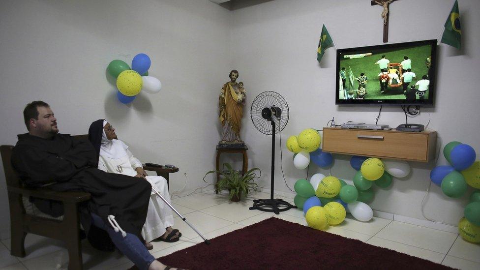 A priest from the Franciscan order and a nun from the enclosed monastery of Imaculada Conceicao watch on television as Brazil's Neymar grimaces as he is carried off the pitch after being injured in the 2014 World Cup quarter-final