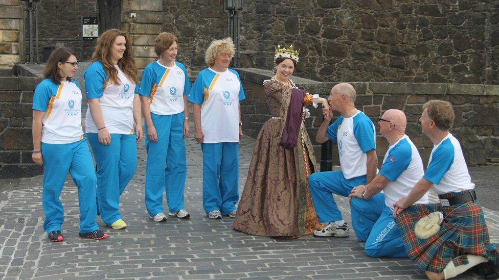 Woman in period costume is kissed on the hand by kneeling baton bearer