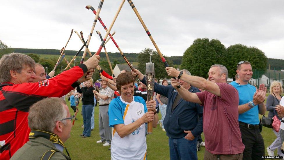 Woman carries baton under an archway formed from crossed shinty sticks