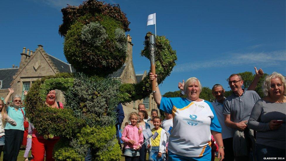 Baton bearer poses next to three metre high shrub pruned to resemble Commonwealth Games mascot Clyde