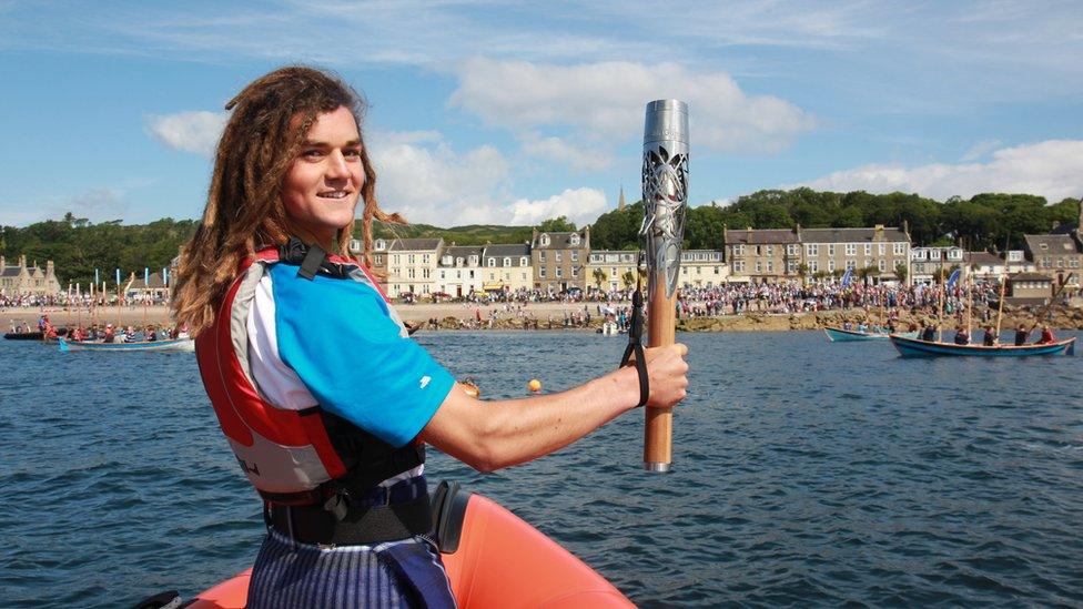 Tristan Levie holds Queen's baton aloft at prow of boat approaching a crowd-lined shore