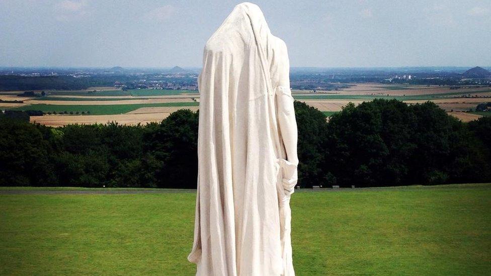 A tall white statue has its head bowed and faces away towards the fields