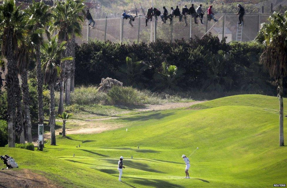 A golfer hits a tee shot as African migrants sit on a border fence in an attempt to cross into Melilla