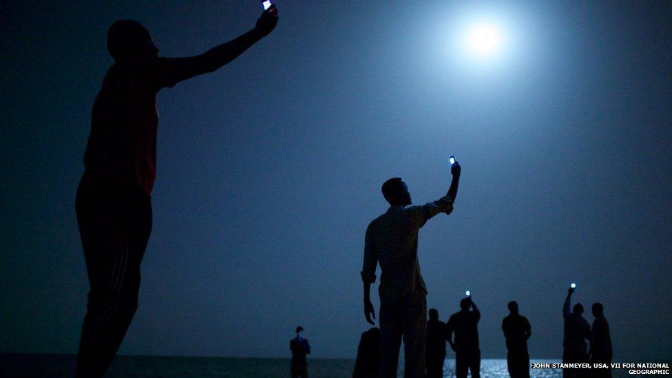 African migrants on the shore of Djibouti city at night, raising their phones in an attempt to capture an inexpensive signal from neighbouring Somalia—a tenuous link to relatives abroad