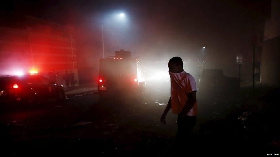 A protester (protestor, demonstrator) walking through the smoke of burning Baltimore buildings set ablaze by rioters as a police armored car drives past during clashes in Baltimore, Maryland 27 April 2015