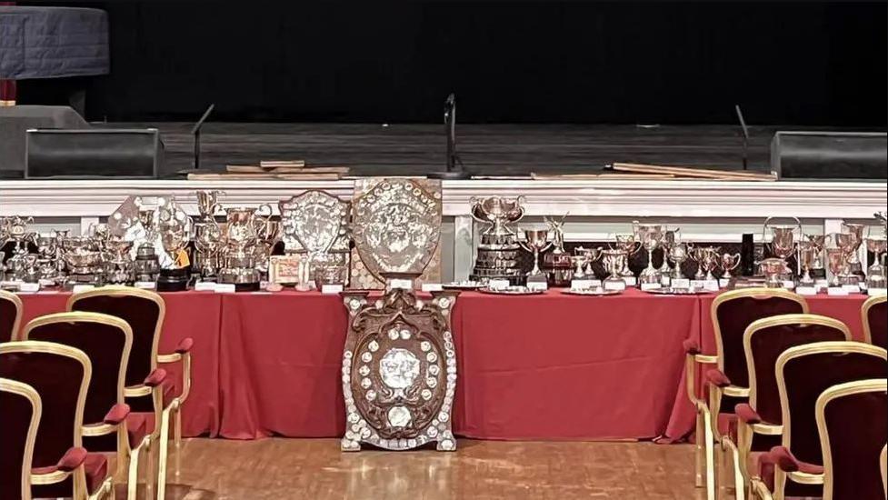 A long red table with multiple brown shields with silver-plated inscriptions. there is also a host of silver trophies on the table, which is in front of a stage. Gold and red chairs have been set out in rows nearby.