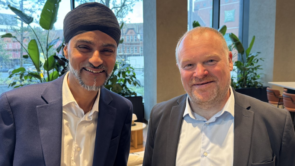 Dav Bansal and Andy Bushell. Dav is a slim man wearing a turban and a blue suit. He has a greying beard and is smiling. Andy is wearing a grey suit. He is smiling at the camera. He has a shaved head and some stubble.