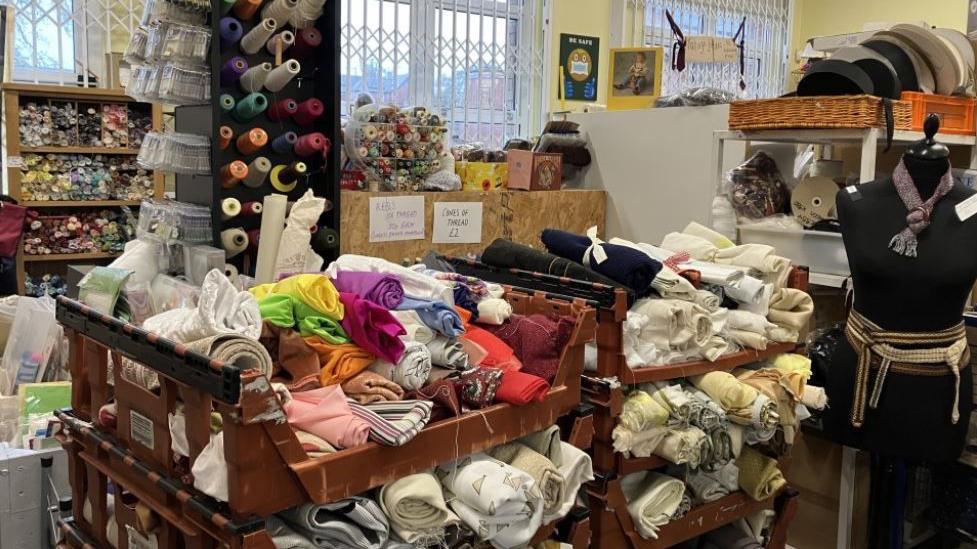 Piles of cloth in brown plastic crates stacked up in a room, with more seen on benches and shelves in the background