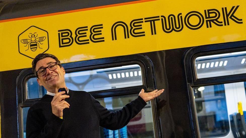 Andy Burnham, in a black jumper holding a microphone, stands in front of a yellow Bee Network vehicle