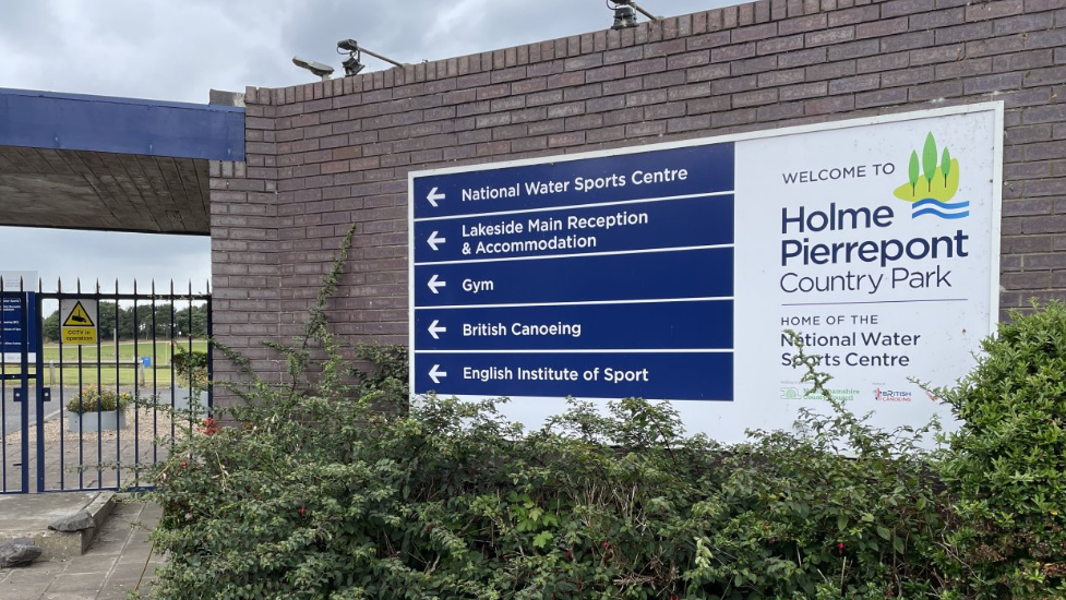 The entrance to Holme Pierrepont Country Park - there is a blue and white sign affixed to a brown brick wall, on the left side of the wall is a blue gate/ A bush is visible in the background