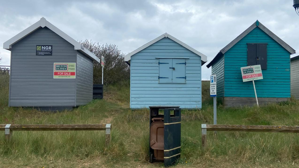Three beach huts are in a line next to each other standing on grass. The one on the right is painted light grey and have a for sale sign on it. The middle one and the one on the right are painted blue and have a window.