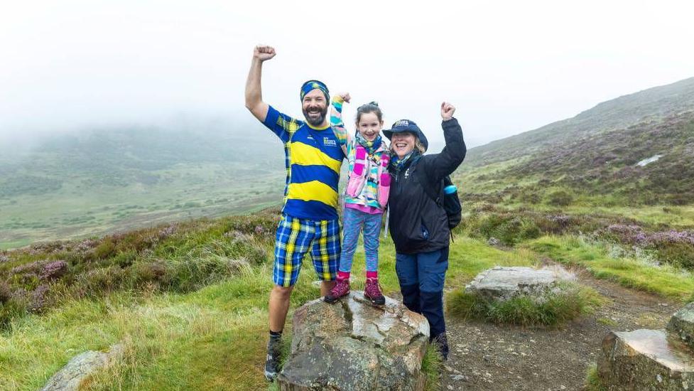 A man, woman and girl hold their hands in the air at the top of a mountain