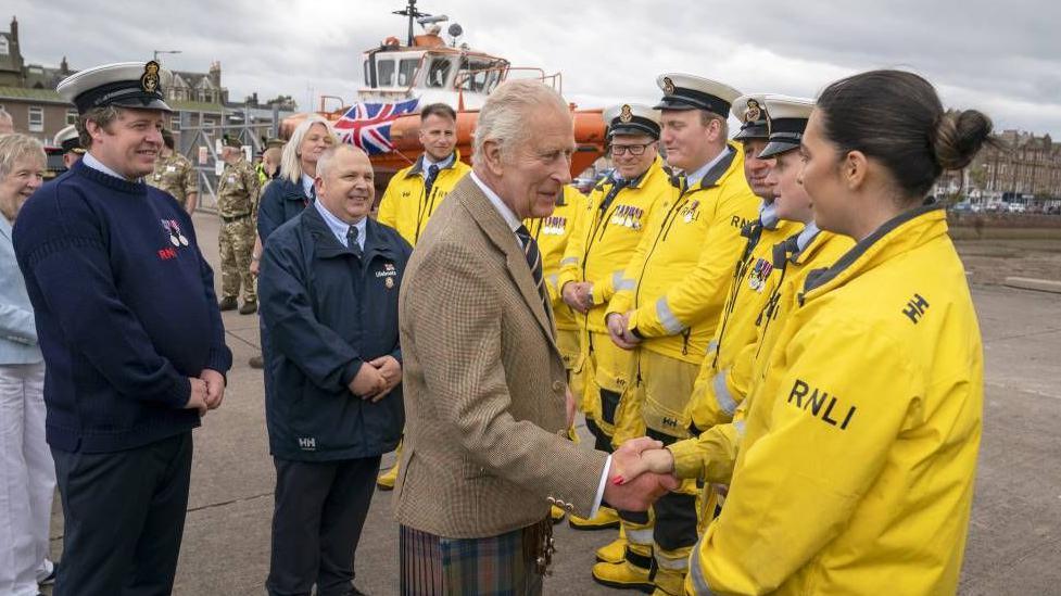 King Charles shakes the hand of a woman wearing an RNLI jacket. She is standing in a line of people with similar yellow jackets. 
