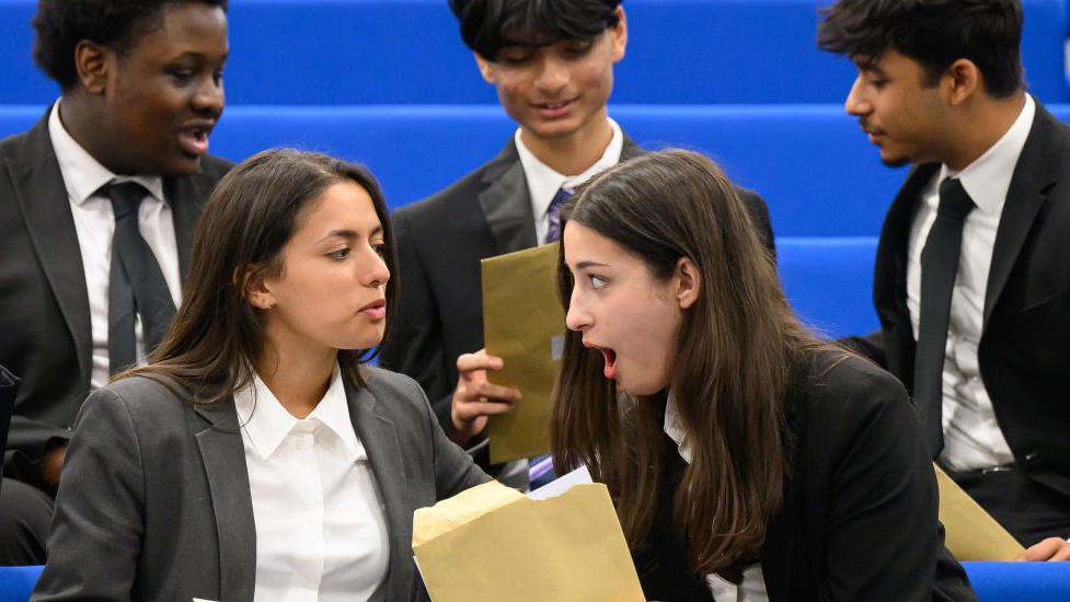 Two female students in uniform that consists of dark blazers and white shirts in conversation with each other. They are holding brown envelopes with level three results inside. Both students have long dark hair and the one on the right looks particularly surprised. There are three male students sitting beside them.