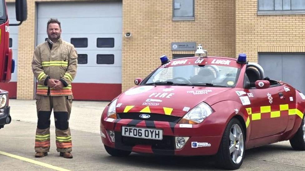 Johnny Smith dressed in a firefighter uniform standing next to his convertible Ford Ka painted outside a fire station.