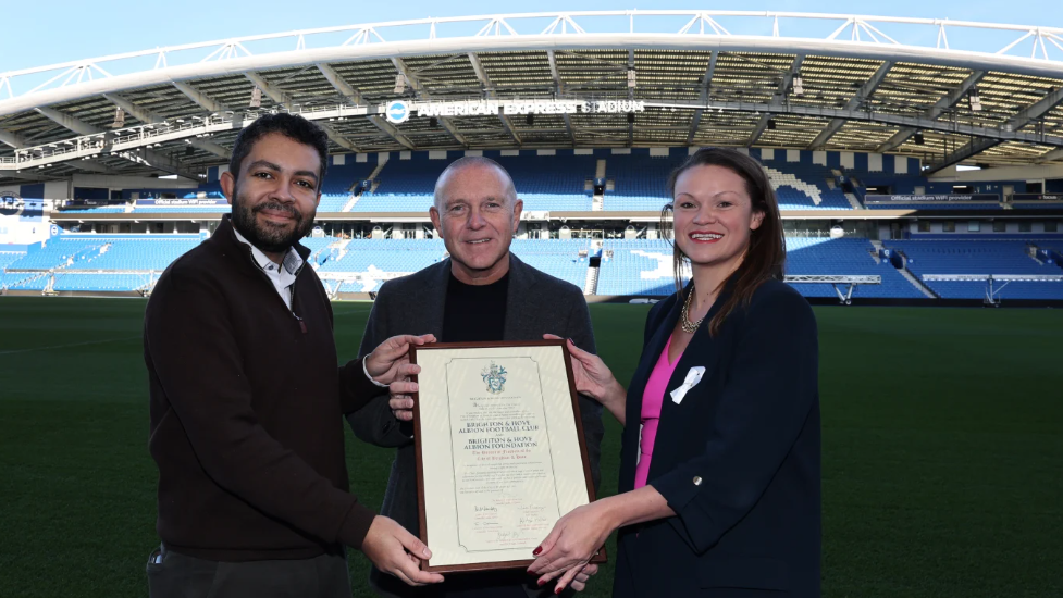 Paul Barber stands between council leader Bella Sankey and cabinet member Leslie Pumm, all holding the Freedom of the City framed certificate on the pitch of the football stadium. The blue seating of the stadium and the green pitch can be seen behind