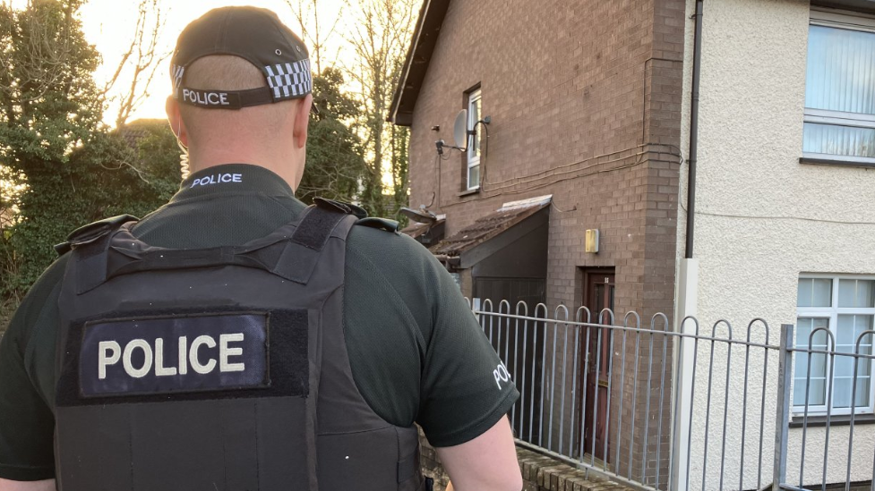 Shows a police officer with their back to the camera near a bricked house with silver railing.
