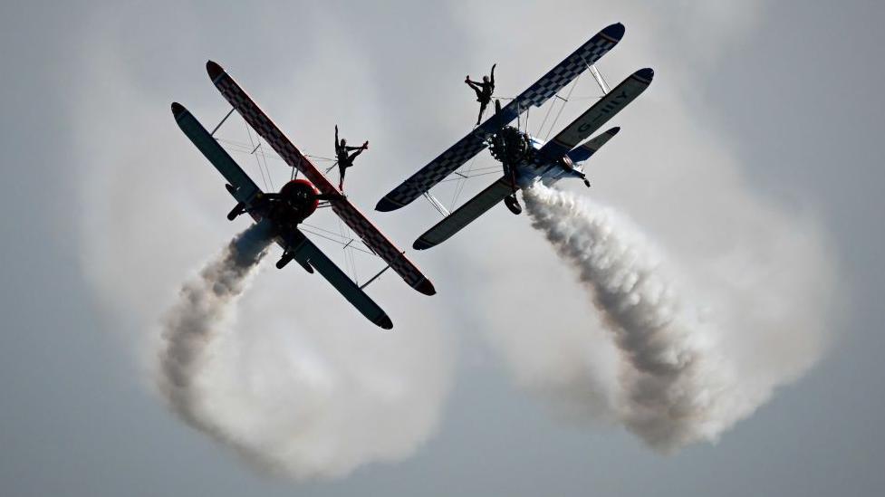 Two planes flying through the air with two aerobatic wing walkers on top, posing with one leg in the air.