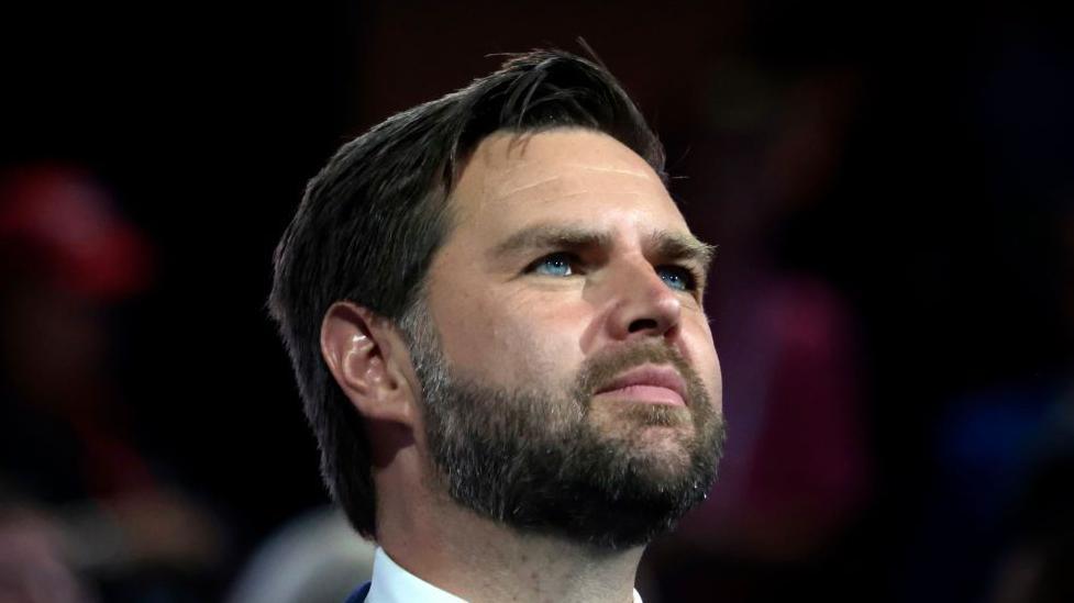 Close up shot of JD Vance looking ahead with piercing blue eyes and dark beard and short hair combed to one side 