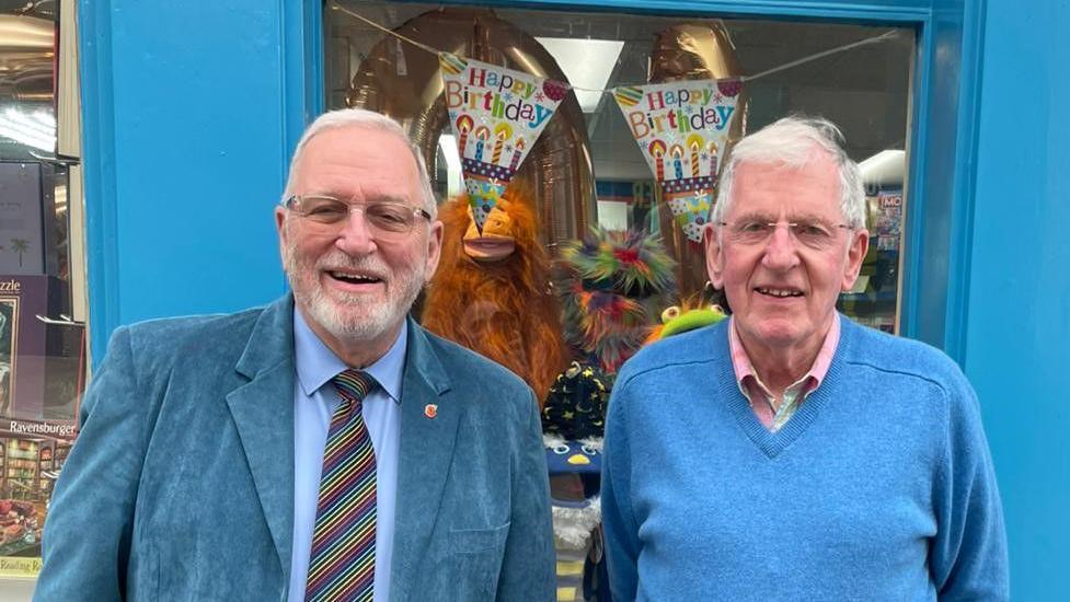 Two men standing outside a shop with a blue-painted façade and a sign reading ‘Holman’s’ above the window. The shop window displays books and colourful decorations, including two large orange balloons shaped like animals wearing party hats and a banner that says ‘Happy Birthday.’