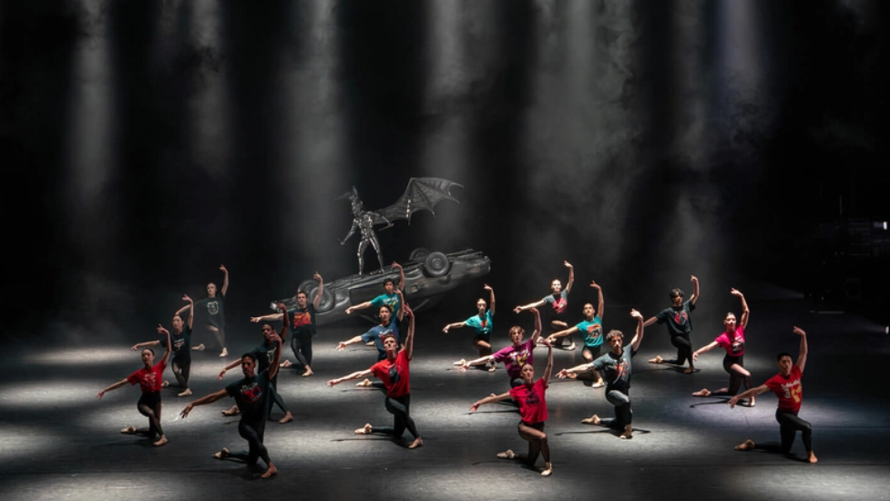 The dance company wearing black tights and colourful shirts on a lit black stage.