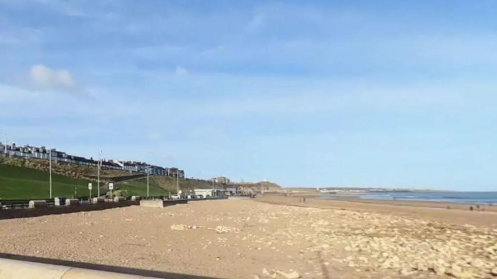 Long view down a stretch stretch of sandy beach under sunny skies. The sea is to the left, and to the right is a promenade with a low wall, with a grassy bank above it topped by a row of terraced houses.
