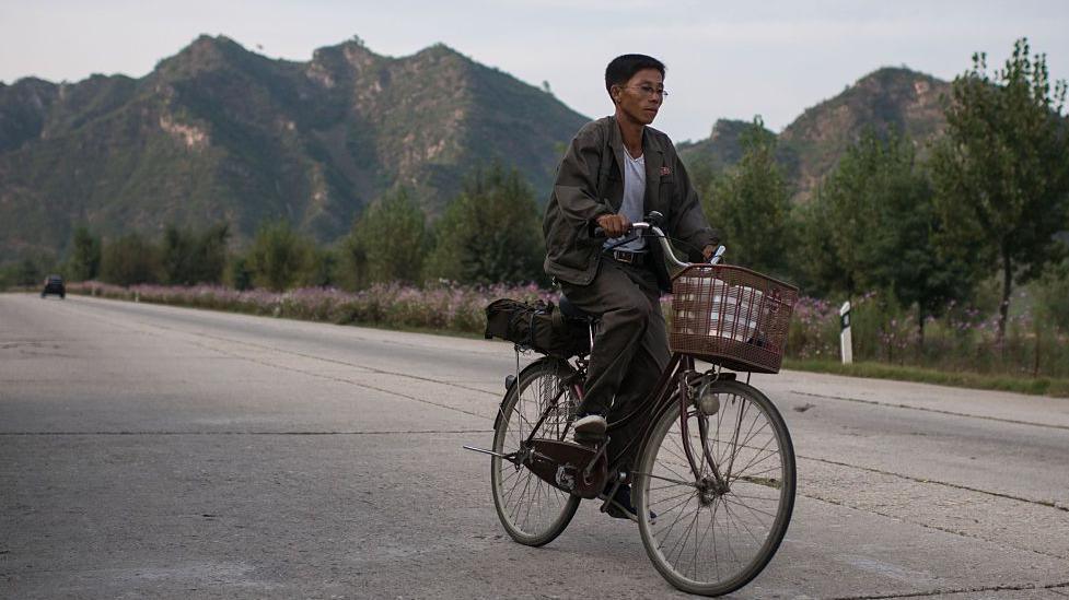 A man in a sweater rides a bicycle down a highway in front of a mountainous background