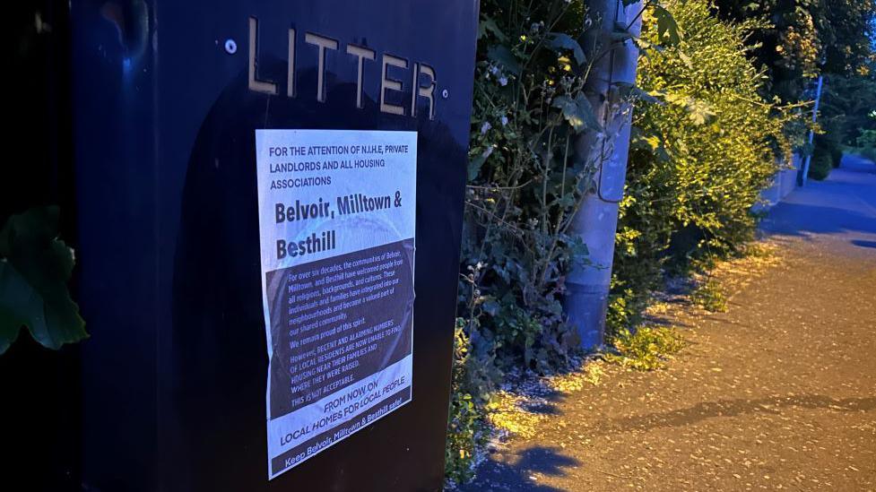 A poster on a black bin. It is brown and white in colour and has text on it which says, for the attention of N.I.H.E, private landlords and all housing associations - Belvoir. Milltown & Besthill - from now on local homes for local people. A street the bottom of a lamppost and some hedges can be seen in the background.