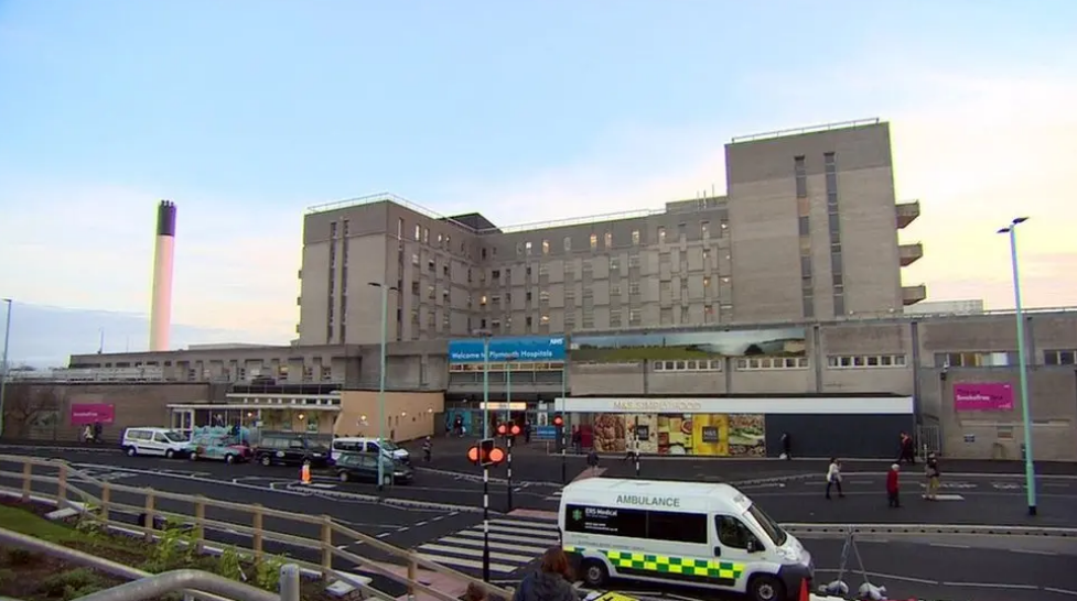 The entrance of the Derriford hospital building from a distance with an ambulance at the front. The building is grey with a pink sign to the right and a blue sign at the entrance. There is a incinerator is behind the building.