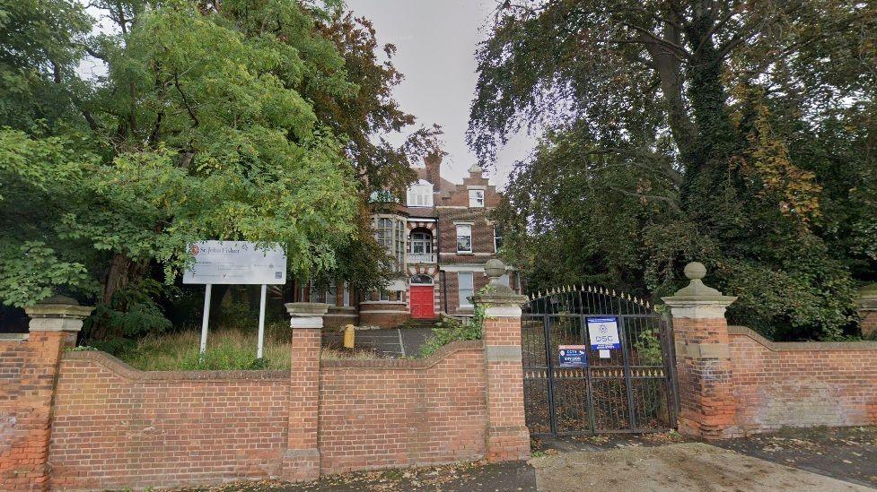 A school building behind a red brick wall and gates. The building is partially hidden by green trees on either side.