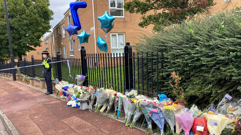 Dozens of bunches of flowers have been laid against a black fence in front of a police cordon. A police officer stands by.