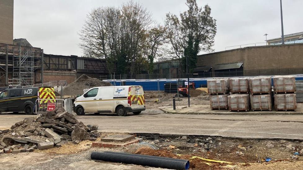 Two small vans with high visibility patterns on them are parked on a concrete area. Pallets piled with construction materials are on the right of the vans. An area in front of them has been dug up and there is a pile of concrete. In the background are two trees, a wall and a building. 