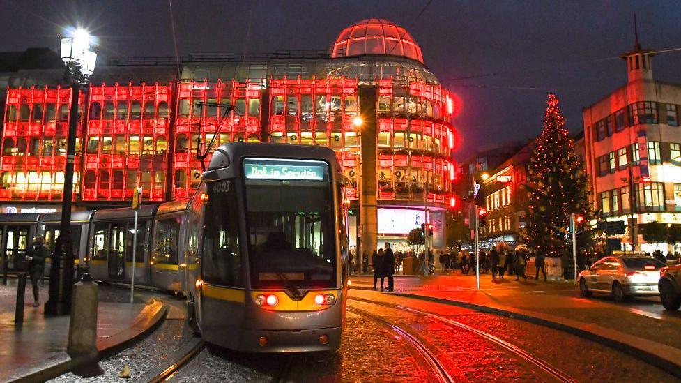 Luas Cross City in Dublin city centre at night time. Red lights shine from St Stephen's Green Shopping Centre and a large Christmas tree and crowds are in the background.