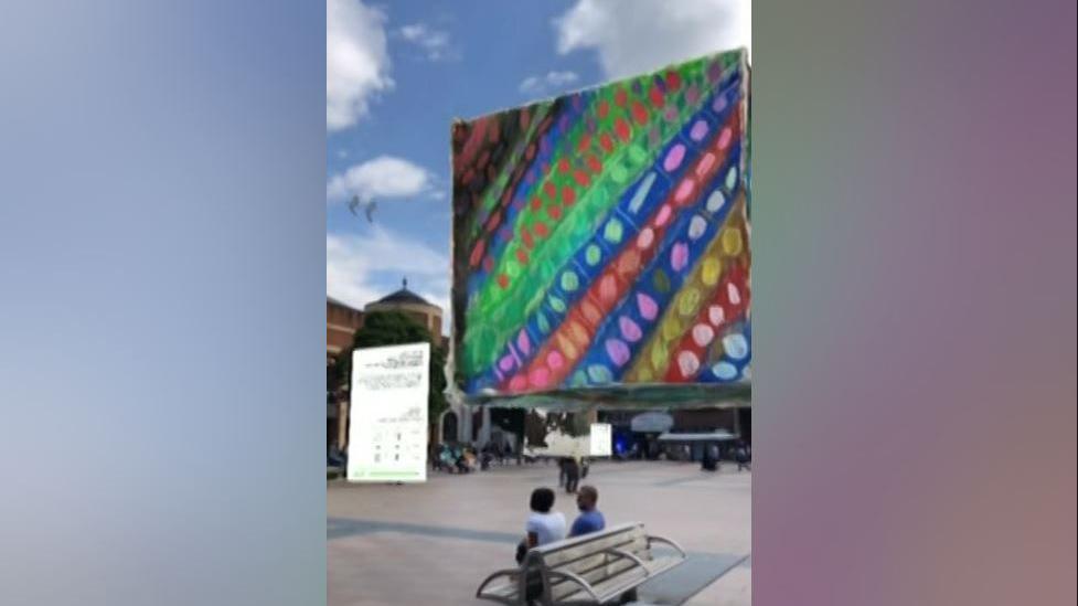 A portrait picture of a large square with colourful strips and dots is floating above Coventry city centre. In the foreground a couple are sat on a bench