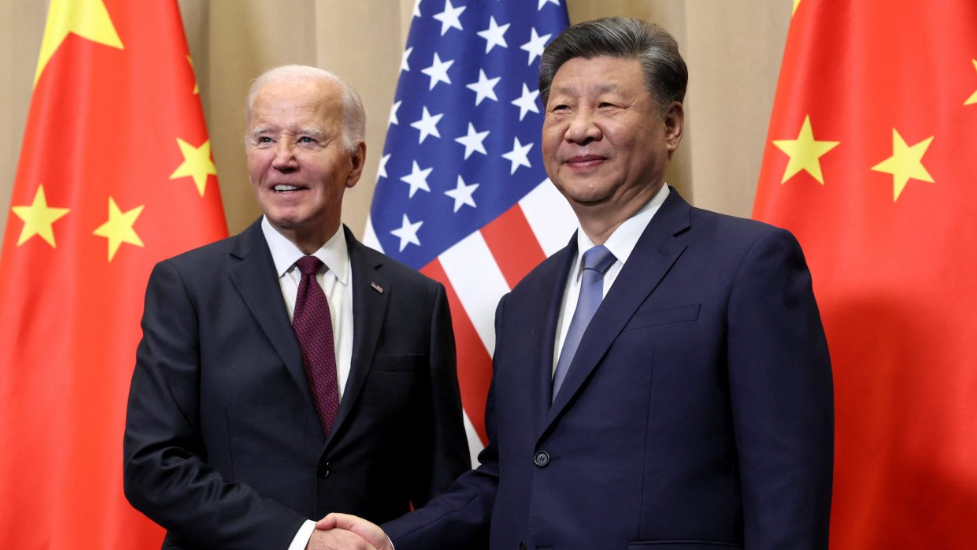 U.S. President Joe Biden and Chinese President Xi Jinping shaking hands in front of a backdrop displaying the flags of the United States and China. Biden is dressed in a dark suit with a white shirt and a burgundy tie, while Xi wears a navy suit paired with a white shirt and a blue tie.