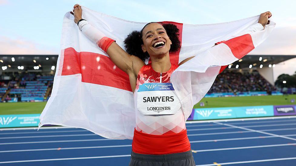 A smiling Jazmin Sawyers in a red and white Team England vest raises her arms to the skies whiles draped in the England flag
