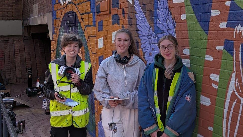 Three women stand in front of a graffiti wall. Two of them are wearing high visibility jackets. The wall is covered in colourful street art