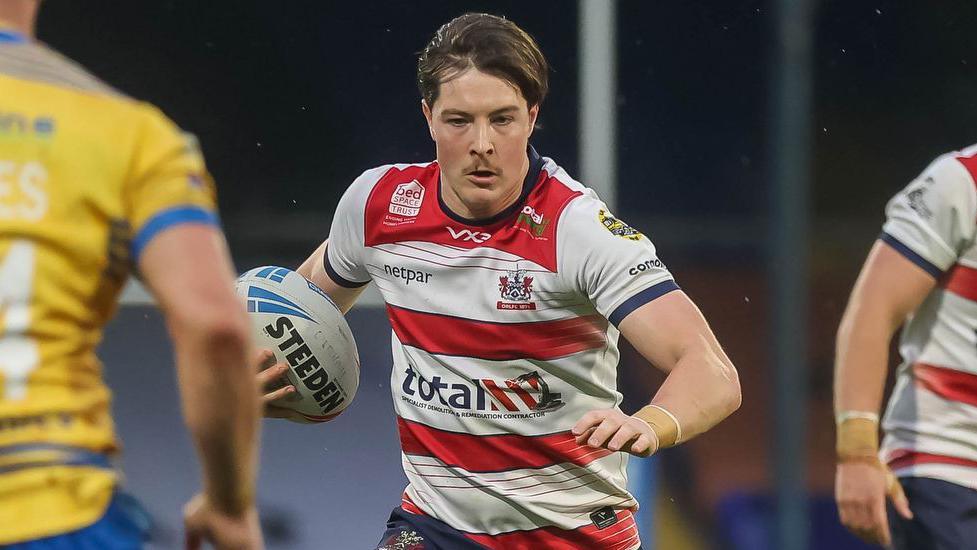 Riley Dean has short dark hair and is running with the rugby ball in one hand. He is wearing a shirt which has red and white hoops.